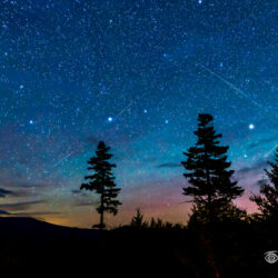 Star-filled night sky with several Perseid meteors above the silhouette of Mount Katahdin and evergreen trees in Maine.